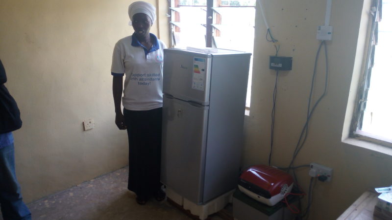Aminichi Edwards, a Junior Community Health Extension Worker (JCHEW) at the Garaha Primary Healthcare Centre in Garaha Mojili, stands beside one of the two fridge-freezers provided the clinic. She says that, compared to before when the hospital had no such equipment, drugs and vaccines such as the Hepatitis B Vaccine can now be stocked and thus readily available for administration to newly born babies